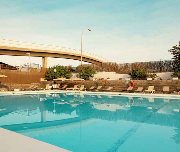 Ulkona oleva uima-allas, jonka ympärillä on aurinkotuoleja ja aurinkovarjoja sekä kasveja ja muuri. (Tekoälyn luoma teksti)