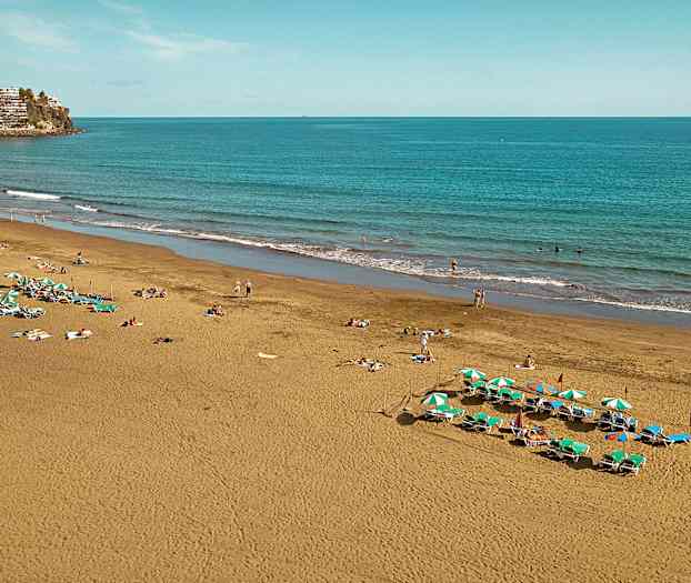 Kuvassa hiekkaranta meren äärellä, jossa on aurinkotuoleja ja varjoja sekä ihmisiä nauttimassa rannasta. (Tekoälyn luoma teksti)