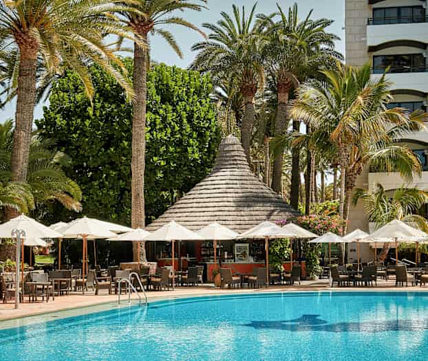 Ulkoalue, jossa on aurinkovarjoja altaan edessä ja rakennus palmuineen taustalla. (Tekoälyn luoma teksti)