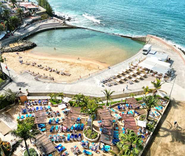 Ilmakuva rannasta, jossa on aurinkotuoleja ja aurinkovarjoja, ympäröity palmuilla ja kävelytiellä. (Tekoälyn luoma teksti)