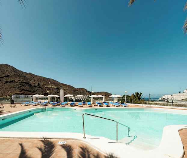 Ulkouima-allas, jossa aurinkotuoleja ja aurinkovarjoja, taustalla vuoria ja kirkas sininen taivas. (Tekoälyn luoma teksti)