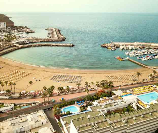 Ilmakuva rannasta, jossa on aurinkovarjoja ja aurinkotuoleja, ympäröivä satama veneineen ja läheisiä rakennuksia. (Tekoälyn luoma teksti)