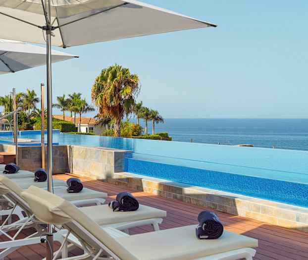 Aurinkotuoleja ja varjoja infinity-altaan äärellä, taustalla näkyy meri. (Tekoälyn luoma teksti)