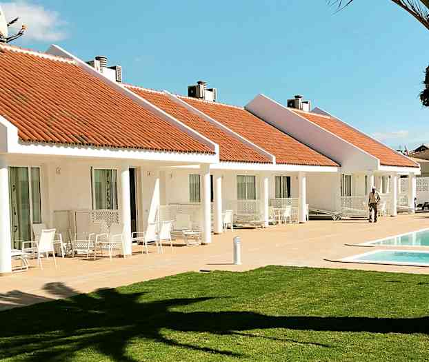 Valkoiset bungalowit oransseilla katoilla ja uima-allas edessä. Nurmikko etualalla. (Tekoälyn luoma teksti)