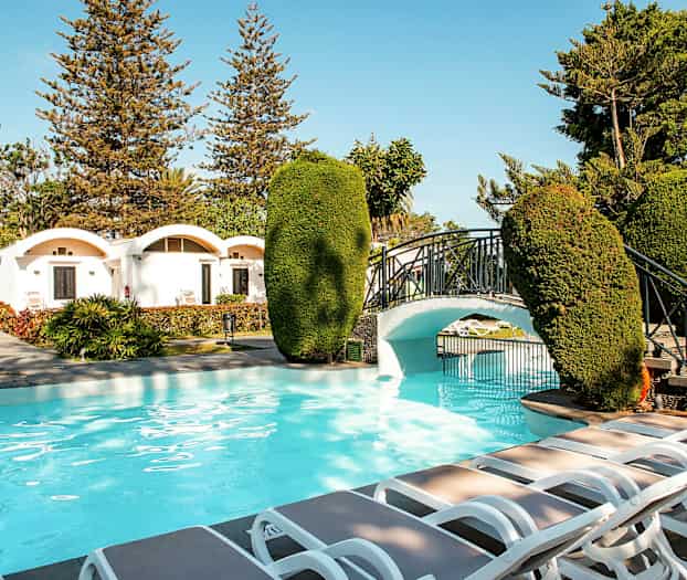 Ulkoallas, jossa on tilava ympäristö, aurinkotuoleja ja pieni silta puutarhassa. (Tekoälyn luoma teksti)