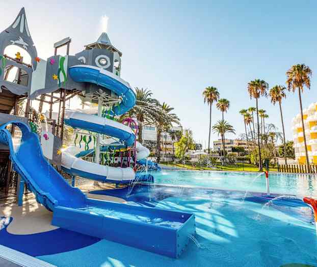 Vesipuisto, jossa vesiliukumäkiä ja leikkipaikka, ympäröity palmuilla ja hotellirakennuksilla. (Tekoälyn luoma teksti)