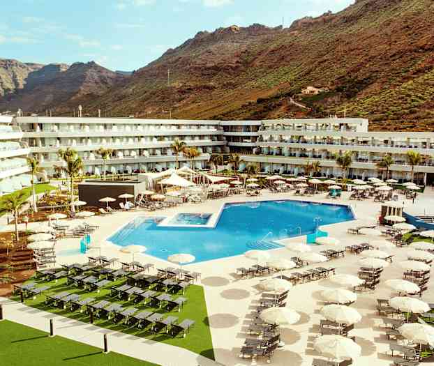 Hotellialue, jossa suuri ulkoallas, aurinkotuoleja ja varjoja, ympärillä rakennuksia ja vuoria taustalla. (Tekoälyn luoma teksti)