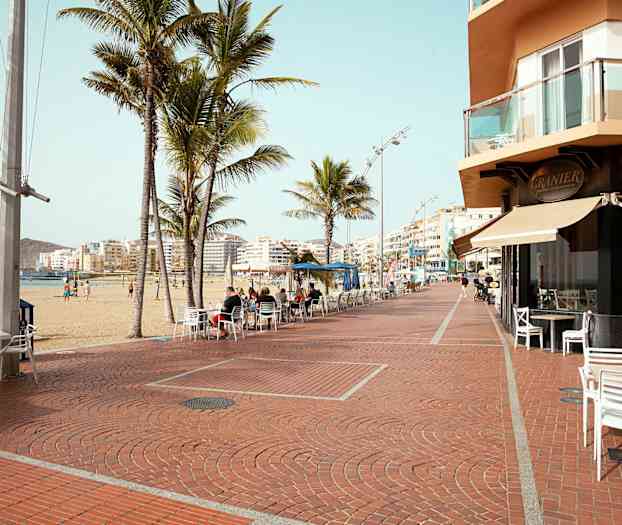 Rantakatu, jolla on palmuja, kahvilatuoleja ja rakennuksia taustalla. (Tekoälyn luoma teksti)