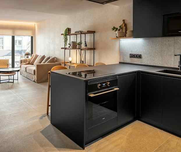 Kuva modernista olohuoneesta ja keittiöstä, jossa on tyylikäs sisustus. Näkyy harmaa keittiösaareke ja sohva taustalla. (Tekoälyn luoma teksti)