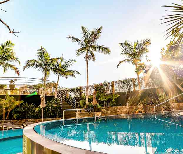 Kirkas uima-allas, palmuja ja ulkoilma-alue taustalla aurinkoisella taivaalla. (Tekoälyn luoma teksti)