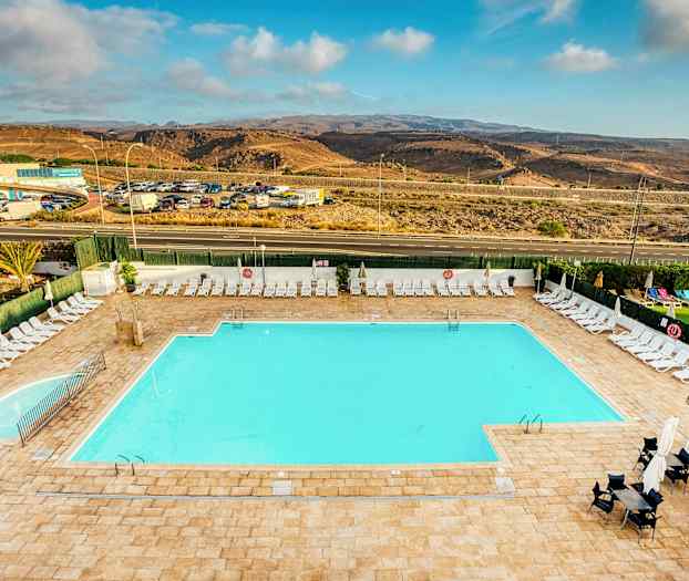 Suuri ulkouima-allas, jota ympäröivät aurinkotuolit ja aurinkovarjot. Taustalla näkyy tie ja kukkulanmuotoista maisemaa. (Tekoälyn luoma teksti)