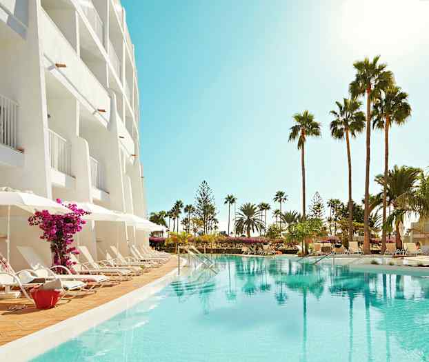 Hotellin julkisivu parvekkeineen, aurinkotuoleja uima-altaan äärellä ja palmuja ympäristössä. (Tekoälyn luoma teksti)