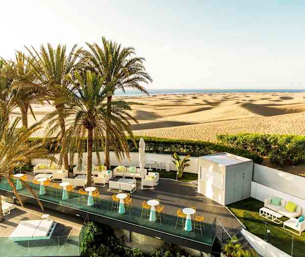 Terassi, jossa on istumapaikkoja ja aurinkovarjoja, autiomaan ja hiekkadyynien edessä, taustalla palmuja. (Tekoälyn luoma teksti)