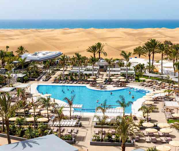 Näkymä hotellin uima-altaalle, jota ympäröivät aurinkotuolit ja aurinkovarjot, sekä taustalla hiekkadyynit ja meri. (Tekoälyn luoma teksti)