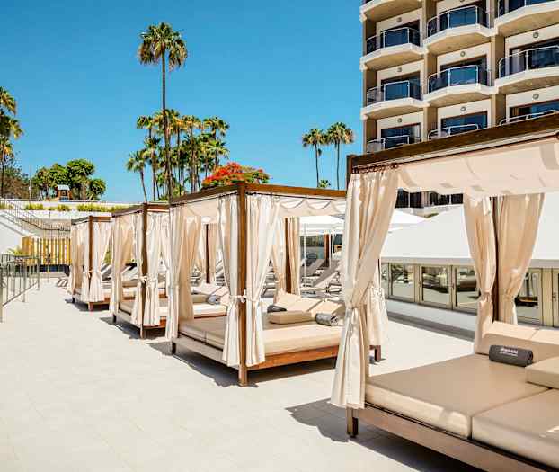 Kuva aurinkotuoleista verhojen kera hotellin uima-altaalla, taustalla palmuja ja rakennus. (Tekoälyn luoma teksti)