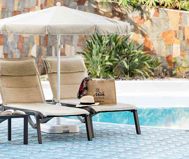 Kaksi aurinkotuolia, joissa on aurinkovarjo uima-altaan äärellä, Juicy-motellilaukku ja hattu. (Tekoälyn luoma teksti)