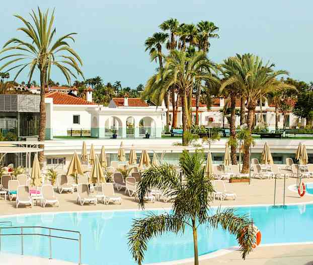 Hotellin allasalue aurinkotuoleilla ja varjoilla, ympäröity palmuilla ja rakennuksilla. (Tekoälyn luoma teksti)