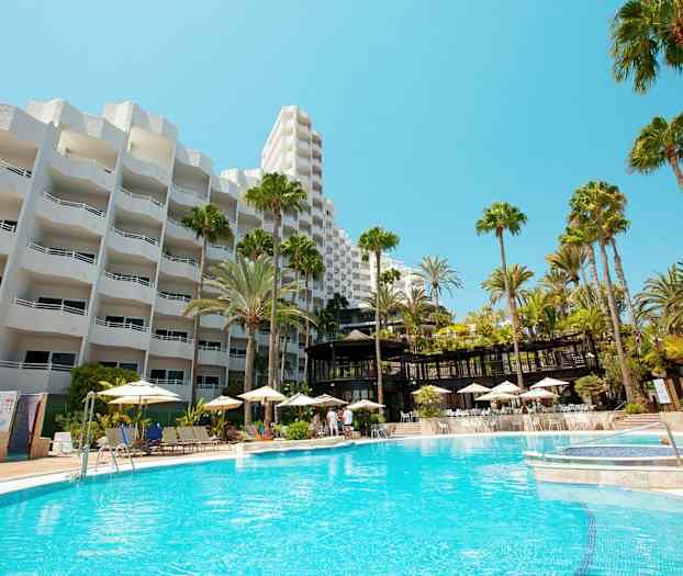 Valkea hotellirakennus ja iso ulkouima-allas, jonka ympärillä on aurinkotuoleja ja varjoja. (Tekoälyn luoma teksti)