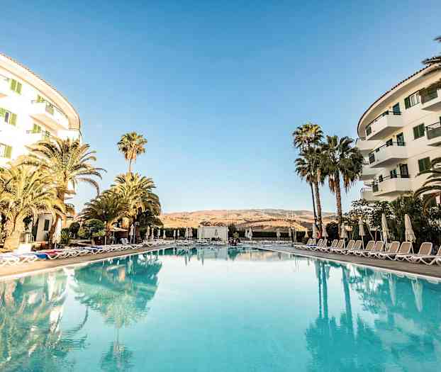 Hotellialue, jossa uima-allas etualalla, ympäröity aurinkotuoleilla ja varjoilla sekä rakennuksilla sivuilla. (Tekoälyn luoma teksti)