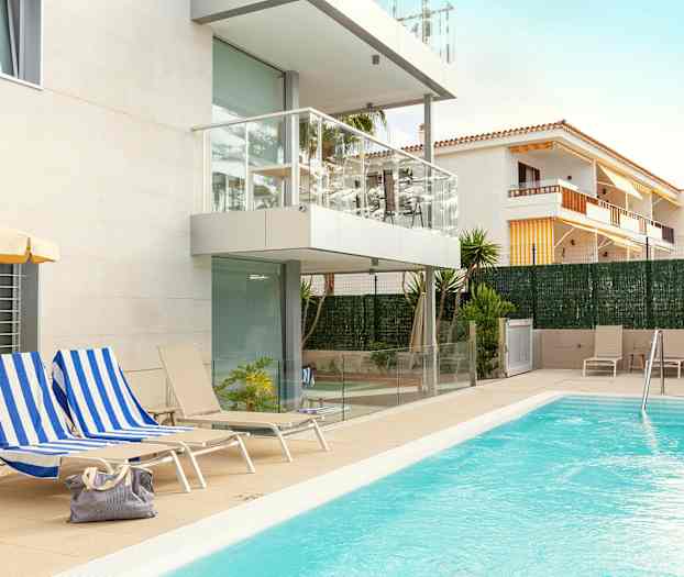 Ulkoallas modernin valkoisen julkisivun ja parvekkeiden vuokratalon vieressä. Aurinkotuoleja ja tuoleja on sijoitettu altaan ympärille, ja yksi laukku on erään aurinkotuolin päällä. (Tekoälyn luoma teksti)