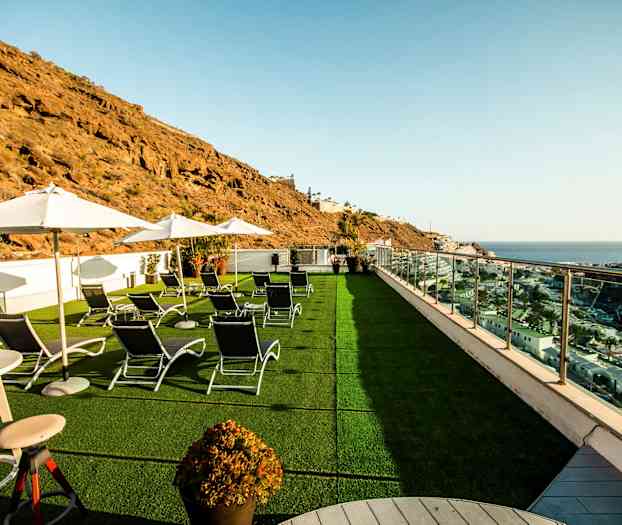 Kuvaus terassista, jolla on aurinkotuoleja ja varjoja, näkymä merelle ja ympäröiviin kallioihin. (Tekoälyn luoma teksti)