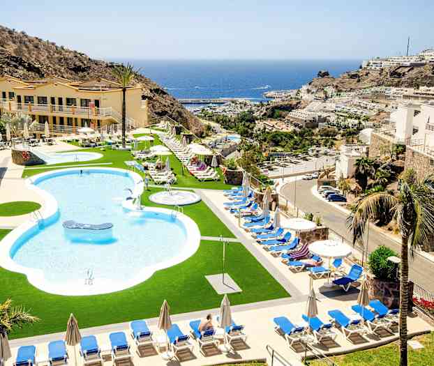 Uima-allasalue aurinkotuoleilla ja -varjoilla, jota ympäröivät rakennukset ja vuoret, taustalla meri. (Tekoälyn luoma teksti)