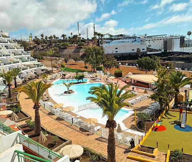 Kuva lomakeskuksesta, jossa on uima-allas ympäröity aurinkotuoleilla ja varjoilla. Etualalla näkyy palmuja ja lasten leikkipaikka. (Tekoälyn luoma teksti)