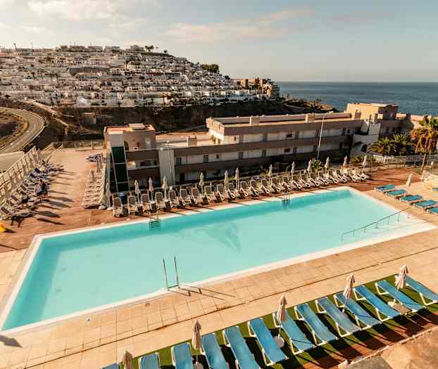 Ulkouima-allas aurinkotuoleineen ja aurinkovarjoineen hotellialueella, merinäköala sekä vuorenrinteen rakennuksia. (Tekoälyn luoma teksti)