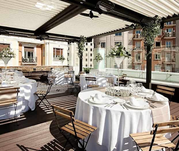 Kuvassa on tyylikäs ulkoalue, jossa on katetut pöydät ja puutuolit katetun pergolan alla. (Tekoälyn luoma teksti)