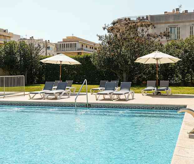 Ulkouima-allas, jossa sinistä vettä, tarjoaa aurinkotuoleja ja varjoja laatoitetulla alueella nurmikon ja vihreiden pensaiden vieressä. (Tekoälyn luoma teksti)