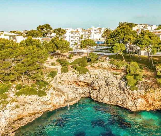 Rannikko, jossa on kirkasta sinistä vettä, kallioita sekä puita ja rakennuksia taustalla. (Tekoälyn luoma teksti)