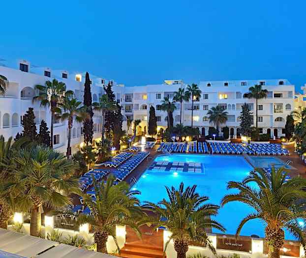 Hotellialue illalla, valaistu uima-allas, ympärillä tuoleja ja vihreitä palmuja. (Tekoälyn luoma teksti)