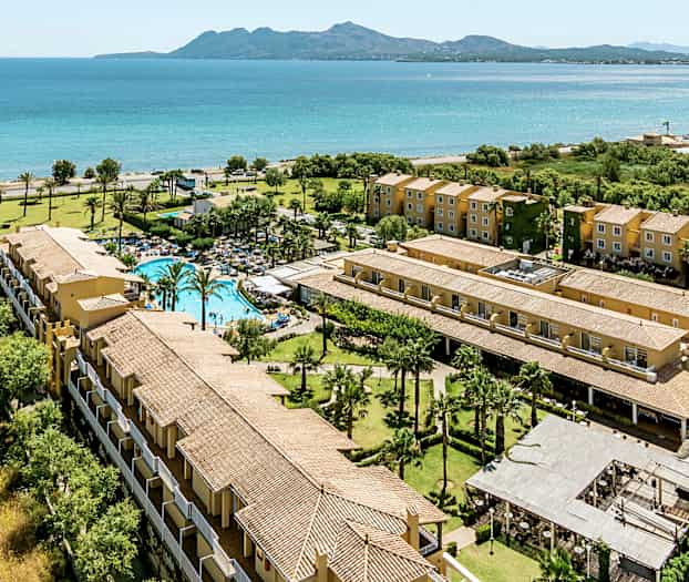 Ilmakuva lomakohteesta meren rannalla, jossa on uima-allas, viheralueita ja moniasuntoisia rakennuksia. (Tekoälyn luoma teksti)