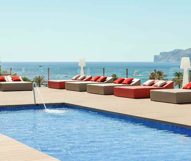 Näkymä uima-altaalle, jossa aurinkovuoteita ja tyynyjä puuterassilla, taustalla meri ja vuori. (Tekoälyn luoma teksti)