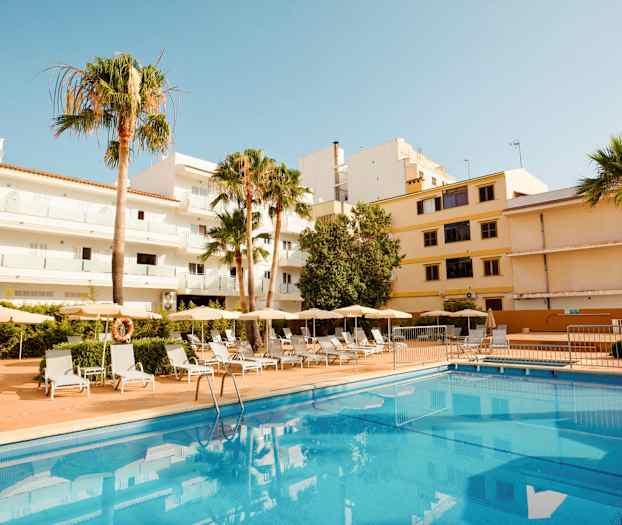 Hotelli ulkopuolelta, jossa uima-allas ja aurinkotuoleja, rakennusten ja palmujen ympäröimänä. (Tekoälyn luoma teksti)