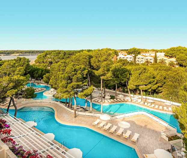 Uima-allasalue aurinkotuoleilla ja -varjoilla, ympärillä puita ja vehreyttä, taustalla lahti ja asuinrakennuksia. (Tekoälyn luoma teksti)