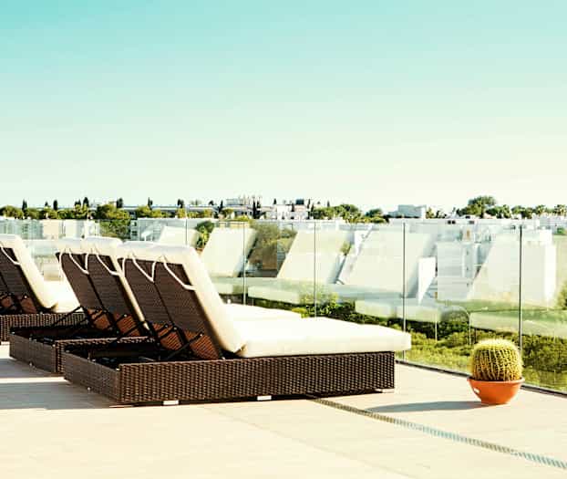 Terassi aurinkotuoleineen ja lasikaiteineen sekä näkymä moderniin asutukseen ja vehreyteen. (Tekoälyn luoma teksti)