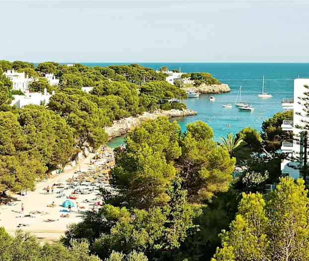 Ranta aurinkotuoleilla ja aurinkovarjoilla vehreiden puiden keskellä, sekä lahti veneineen ja kirkkaan sininen vesi. (Tekoälyn luoma teksti)