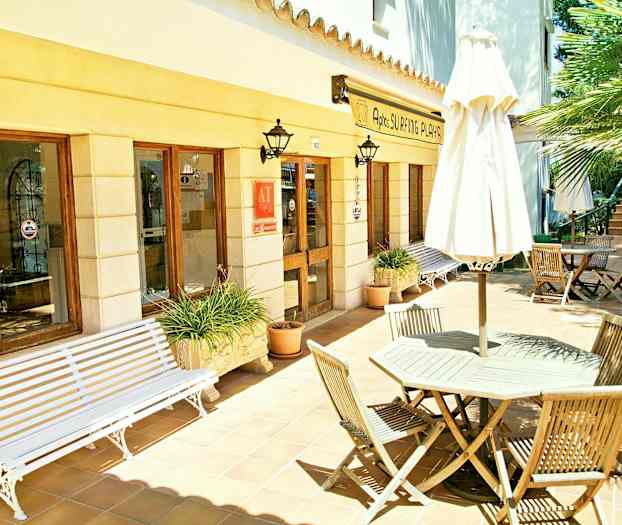 Rakennuksen ulkoterassi, jossa on ikkunoita ja sisäänkäynti. Puuhuonekalut ja aurinkovarjo ovat laattalattialla. (Tekoälyn luoma teksti)