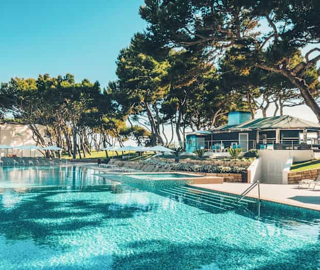 Suuri uima-allas, jota ympäröivät aurinkotuolit ja aurinkovarjot, taustalla rakennus ja puita. (Tekoälyn luoma teksti)