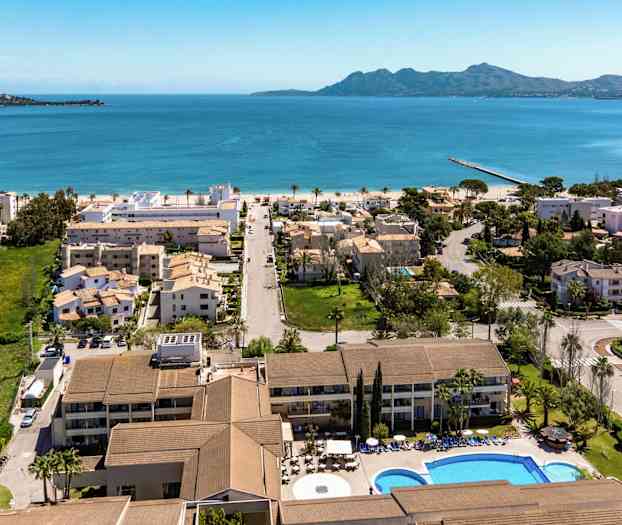 Ilmakuva rannikkokaupungista, jossa hotelli, asuinrakennuksia ja uima-allas etualalla, sininen meri ja vuoret taustalla. (Tekoälyn luoma teksti)