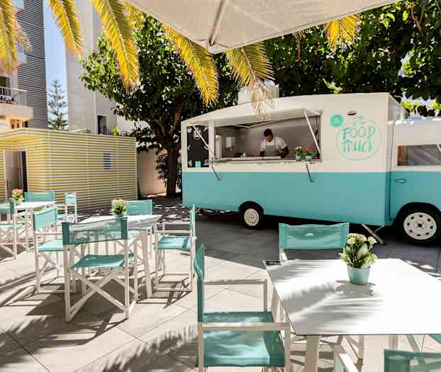 Sinivalkoinen ruokarekka on pysäköity pöytien ja tuolien viereen, ympäröitynä puilla ja aurinkovarjoilla. (Tekoälyn luoma teksti)