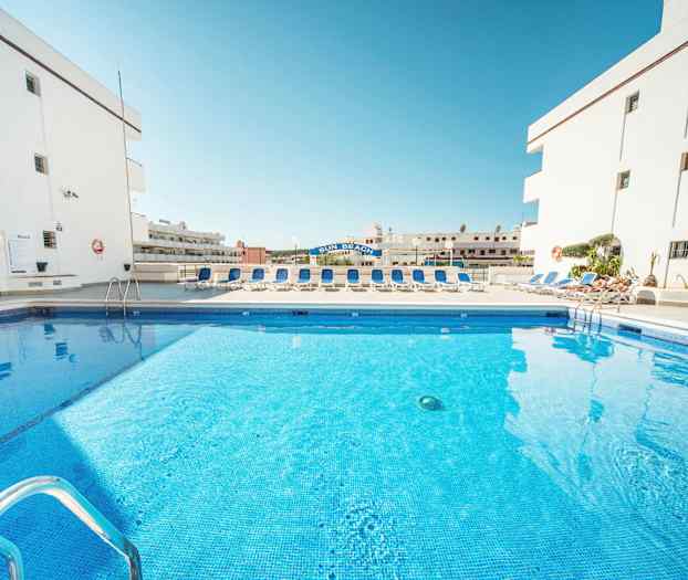 Kuva uima-altaasta, jota ympäröivät aurinkotuolit ja valkoiset rakennukset. (Tekoälyn luoma teksti)