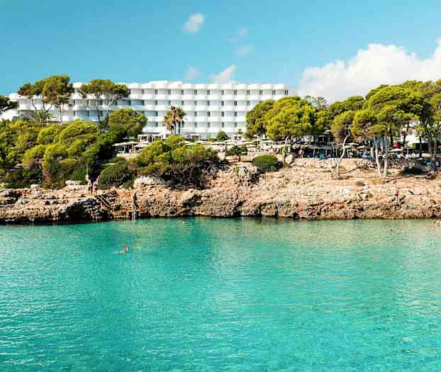 Hotelli, jossa on valkoisia rakennuksia kallioisen rannan takana ja kirkasta sinistä vettä. (Tekoälyn luoma teksti)