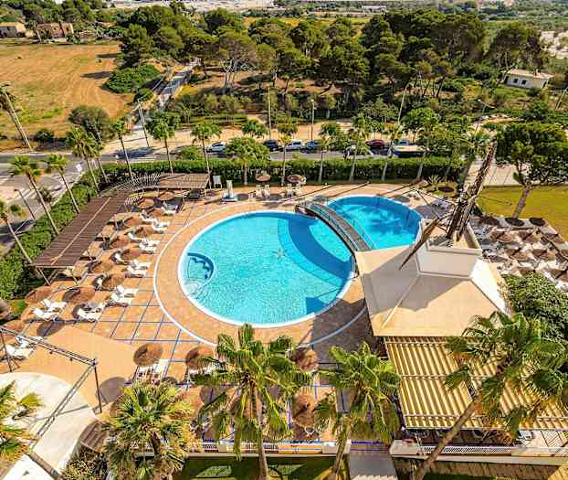 Ilmakuva ulkoalueesta, jossa on kaksi uima-allasta, aurinkotuoleja ja varjoja, vehreyden ympäröimänä. (Tekoälyn luoma teksti)