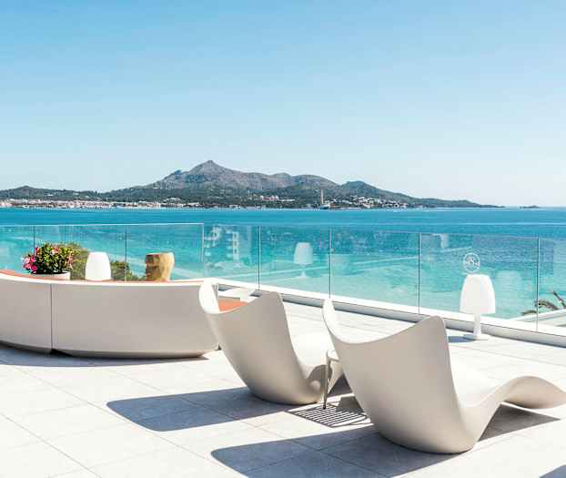 Näkymä terassilta, jossa moderneja istuimia ja lasikaide, meri ja vuoristoinen rannikko taustalla. (Tekoälyn luoma teksti)