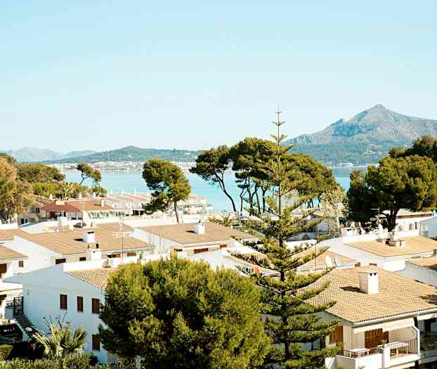 Useita valkoisia taloja tiilikatoilla, ympäröitynä puilla, taustalla vuoristo ja meri. (Tekoälyn luoma teksti)