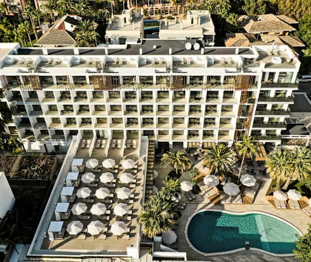 Ilmakuva hotellista, jossa on uima-allas ja suuri terassi, jossa on aurinkovarjoja ja aurinkotuoleja. (Tekoälyn luoma teksti)