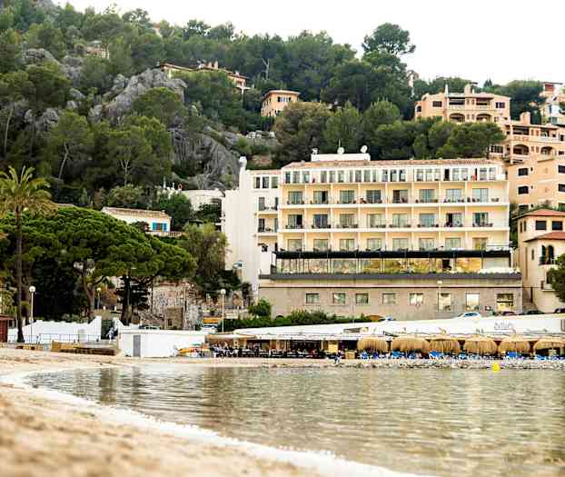 Kuva rannasta, jossa on kirkasta vettä, pieni ravintolarakennus ja monikerroksisia rakennuksia ympäröivien viheralueiden keskellä. (Tekoälyn luoma teksti)