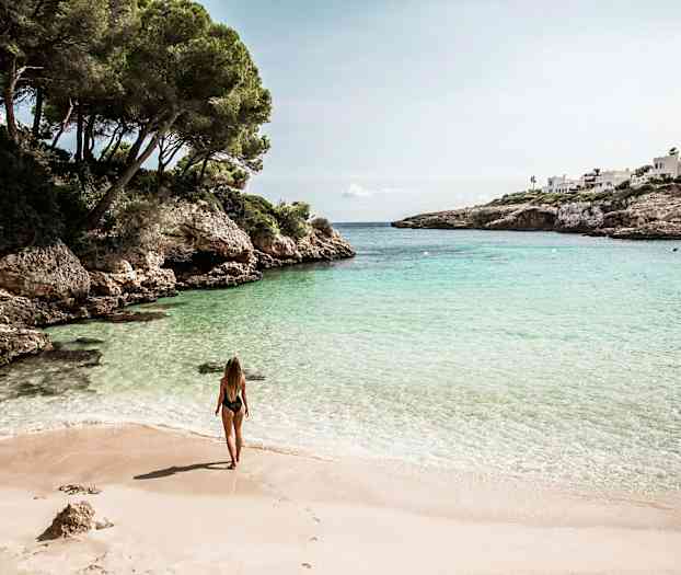 Nainen seisoo hiekkarannalla poukaman äärellä, jossa on kirkasta vettä ja ympärillä kallioita ja vehreyttä. (Tekoälyn luoma teksti)
